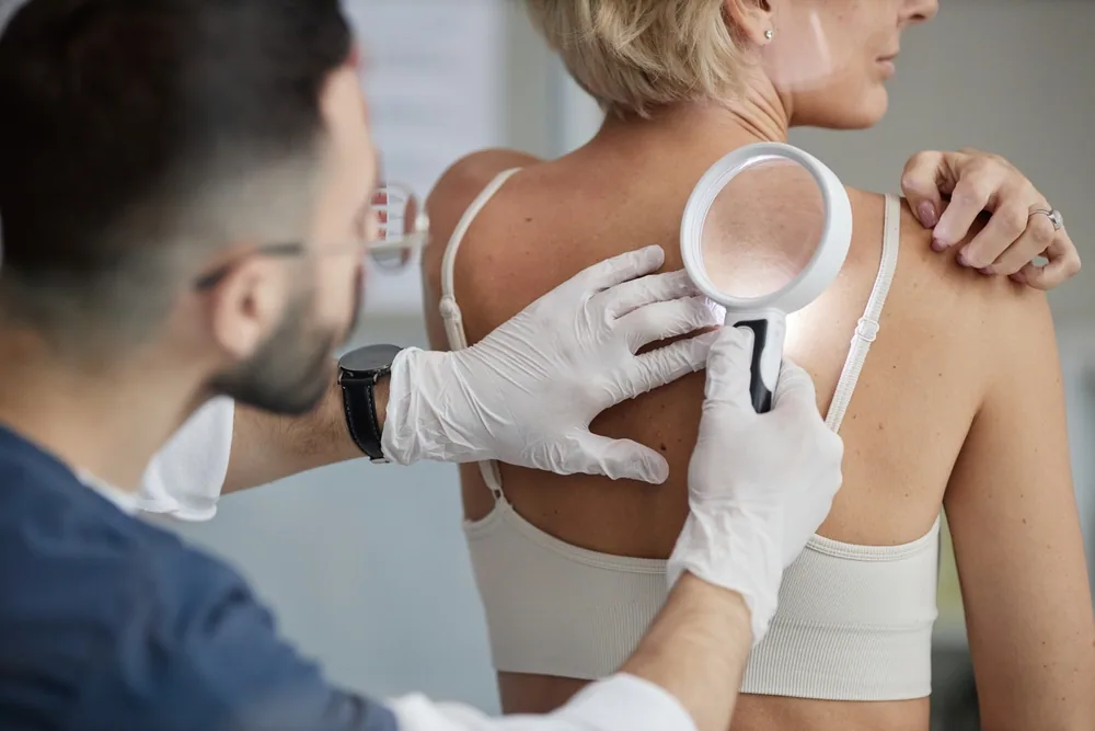 A doctor in a white coat applies cream to a woman's arm, demonstrating a step in a skin self exam how to.