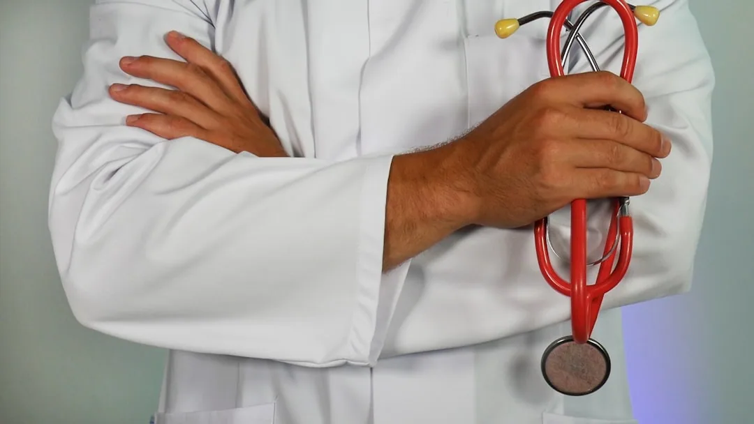 A doctor in a white coat stands with arms crossed, holding a red stethoscope, perhaps considering if Mohs surgery is painful.